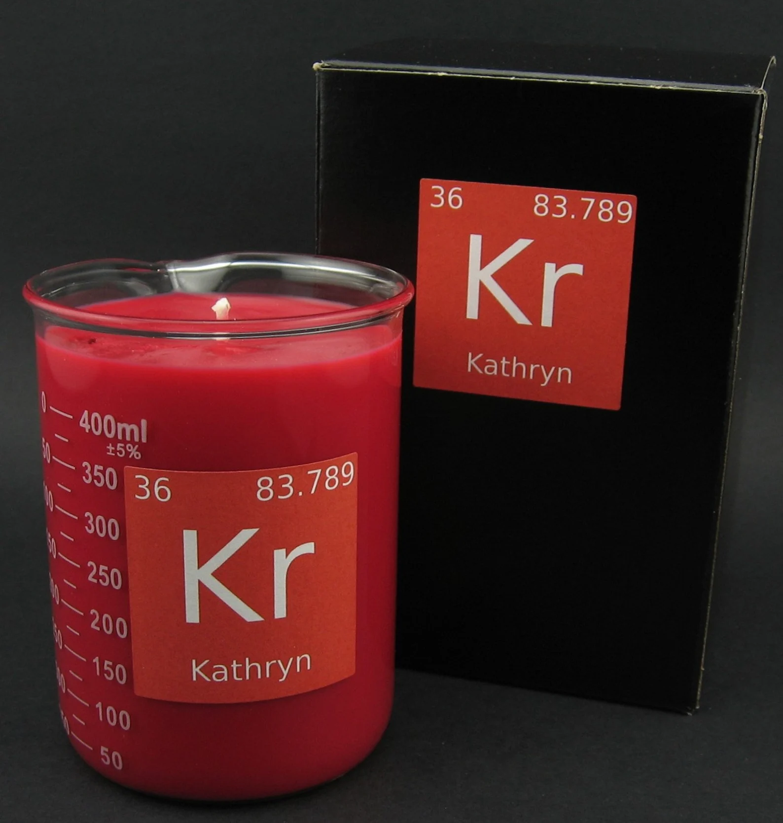 Pink candle in a beaker with a rectangular label that looks like a periodic table entry but reads "Kathryn KR" sits in front of a slightly larger box.