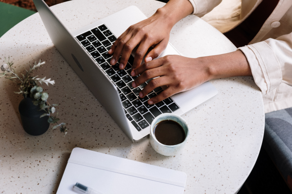 Looking from above, there is a person sitting to the right and you can see their hands typing on a laptop. They are at a small round table with a cup of coffee by them and a succulent plant behind the laptop. There's also a notebook on the table.